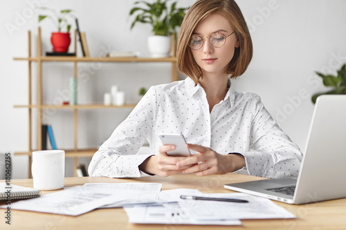 Elegant buisnesswoman dressed formally, surrouded with papers and documents, recieves business message on smart phone, types answer, uses modern electronic gadgets for work and communication photo