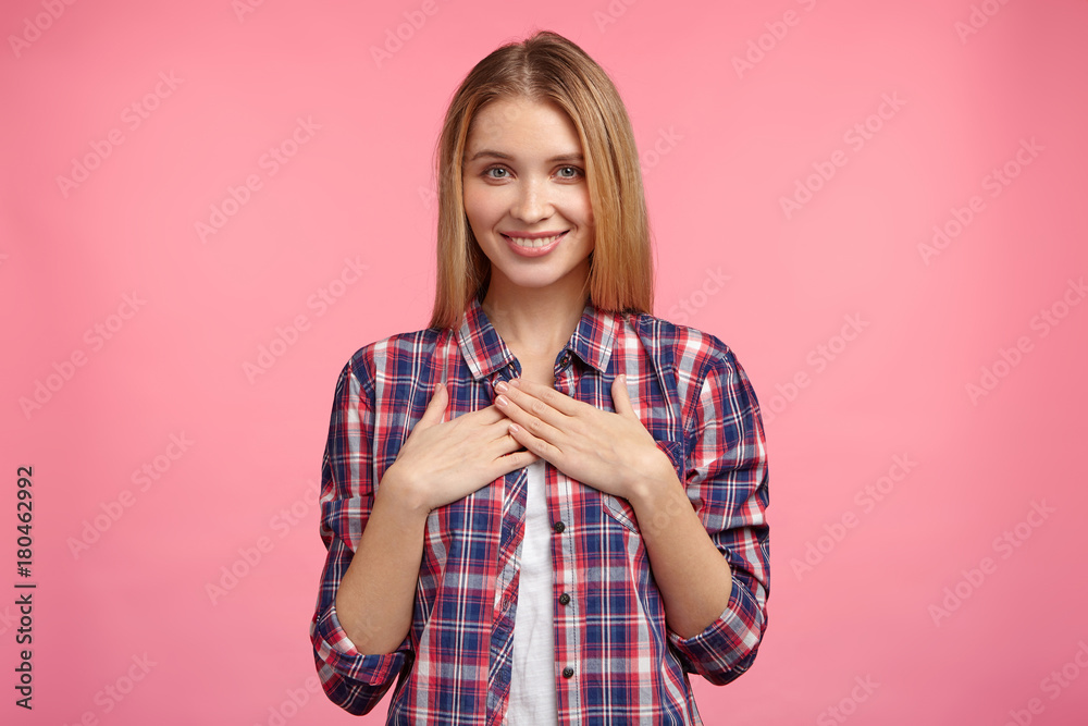 Studio shot of pleasant looking female with appealing smile, keeps hands on chest, demonstrates good feelings and emotions, isolated over pink background. Restful happy student being at home Studio shot of pleasant looking female with appealing smile, keeps hands on chest, demonstrates good feelings and emotions, isolated over pink background. Restful happy student being at home