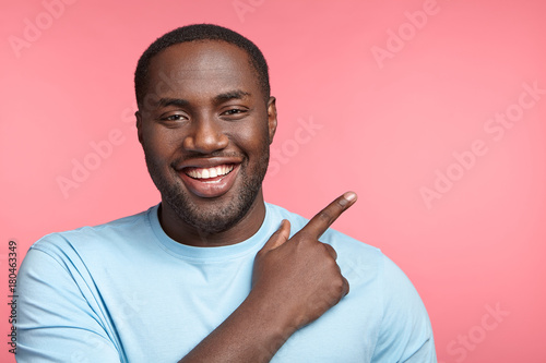African American male grins at camera, indicates at copy space, advertises something. Happy black man points with fore fiinger, has happy expression, isolated over pink background. Look here! photo