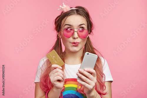 Horizontal portrait of female chick has dreamful expression, imagines new clothes, going do online shopping, holds modern cell phone and plastic card, isolated over pink background with copy space photo