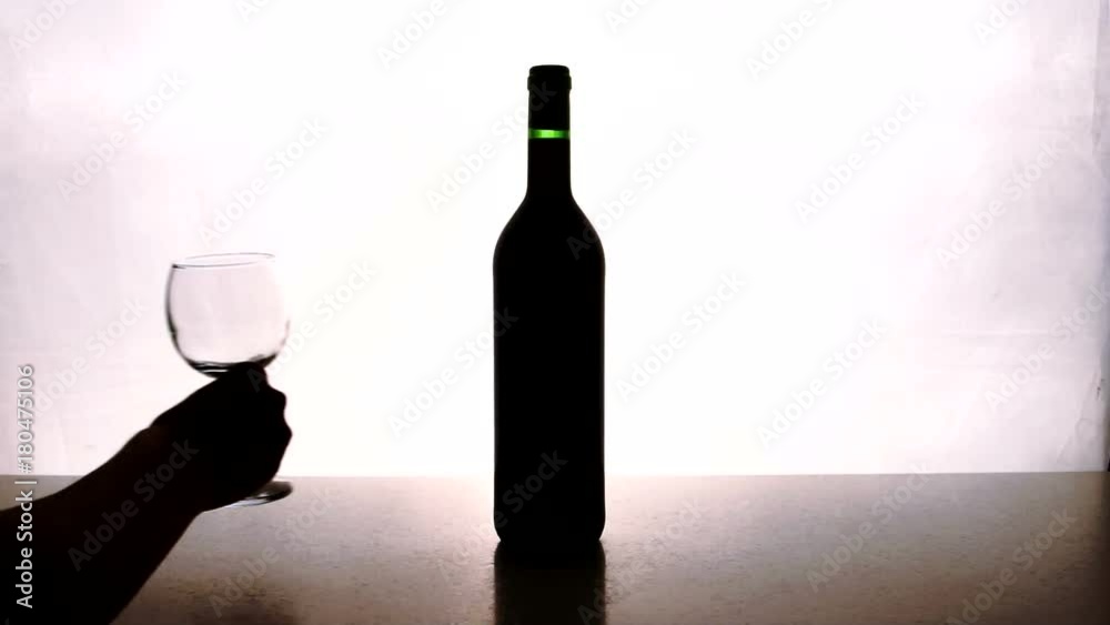 People put on table bottle of wine and two glasses indoor. Man sets bottle on desk with alcoholic drink, holding it by neck confidently then putting glassy transparent glasses in turn. Objects are