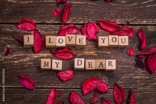 I love you my dear. Valentines card. Small wooden letters on wood cubes standing on dark rustic vintage background. Red and violet pink petals around at the sides. Greetings postage anonymous message.