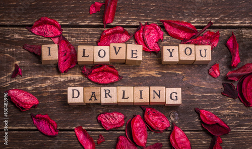 I love you darling. Valentines card. Small wooden letters on wood cubes standing on dark rustic vintage background. Red and violet pink petals around at the sides. Greetings postage anonymous message.