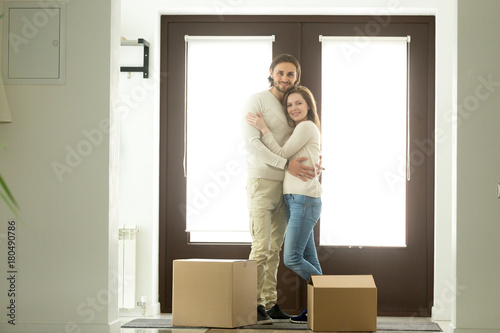 Happy young couple hugging looking at camera moving into new home, smiling homeowners embracing in hallway standing against door near cardboard boxes, affordable mortgage loan, buying house, portrait