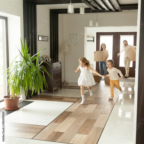 Playful happy kids running into new big own beautiful house, family moving in day concept, excited children exploring home interior having fun together, parents holding cardboard boxes at background
