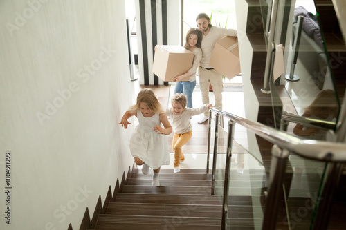 Happy children going upstairs inside two story big house, excited kids having fun stepping walking up stairs running to their rooms while parents holding boxes, family moving in relocating new home
