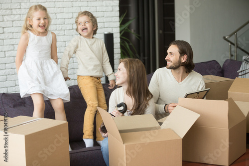 Happy excited young family with kids playing in new home living room on couch, father mother and children having fun on sofa unpacking cardboard boxes together, moving into house relocation concept