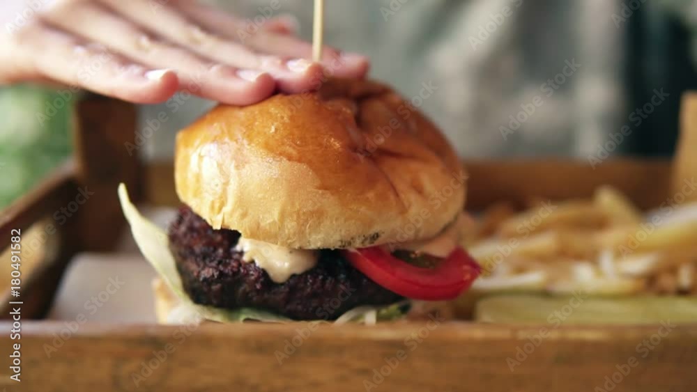 Tasty burger with french fries on a wooden tray. Female hands touching it to start eating. Slowmotion shot Tasty burger with french fries on a wooden tray. Female hands touching it to start eating. Slowmotion shot