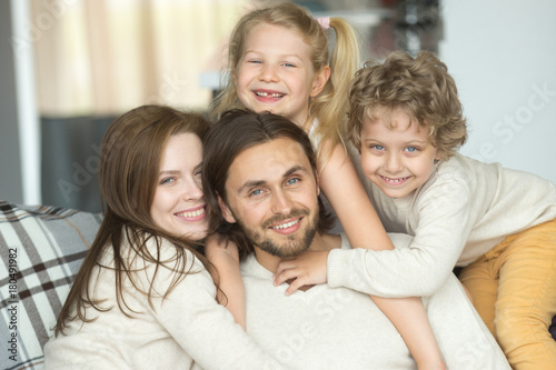 Portrait of happy cheerful young family bonding together, smiling kids embracing parents indoors, son and daughter with mother hugging dad posing looking at camera at home, celebrating fathers day