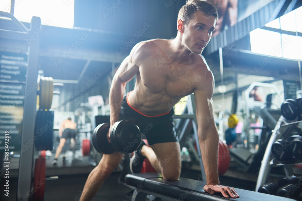 Motivational portrait of handsome muscular man with bare chest working out with dumbbells on bench in modern gym, copy space Motivational portrait of handsome muscular man with bare chest working out with dumbbells on bench in modern gym, copy space