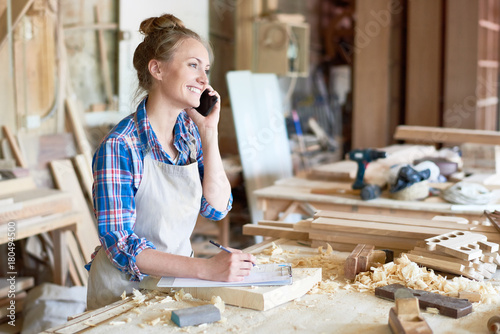 Portrait of modern young woman speaking by phone with client smiling and making notes in woodworking shop photo