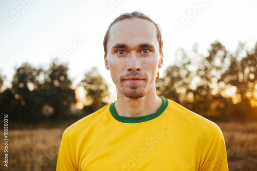 Fototapeta Naklejka Na Ścianę i Meble -  Attractive serious young European man, standing in a meadow, wearing yellow sportswear looking at camera on sunset flare