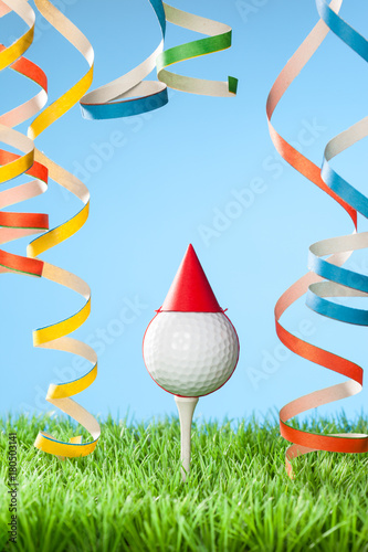 Concept for celebration events at a golf course or for a golf enthusiast with golf ball on tee wearing party hat and blow-out streamers coming down from top