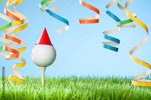 Concept for celebration events at a golf course or for a golf enthusiast with golf ball on tee wearing party hat and blow-out streamers coming down from top