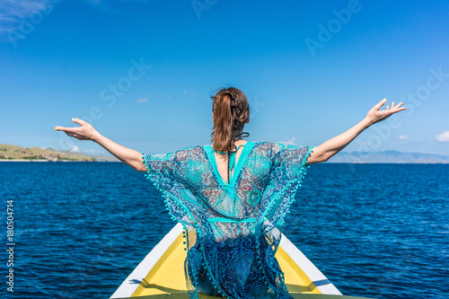 Rear view of a young woman sitting with outstretched arms against a clear blue sky while enjoying summer vacation in Flores Island, Indonesia