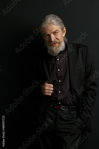 Stylish aged man with lonf hair and beard standing in studio against black wall. Portrait of very old wrinkled male.