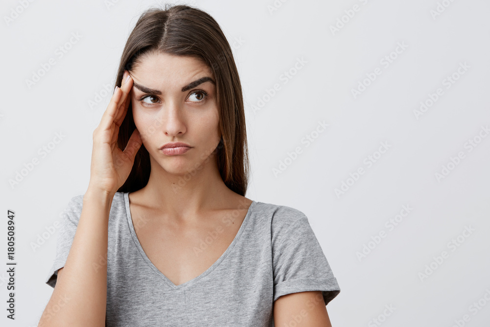 Close up of young handsome serious caucasian girl with long brown hair in stylish gray t-shirt touching forehead with hand, looking aside with tired face expression after long day in university. Close up of young handsome serious caucasian girl with long brown hair in stylish gray t-shirt touching forehead with hand, looking aside with tired face expression after long day in university.