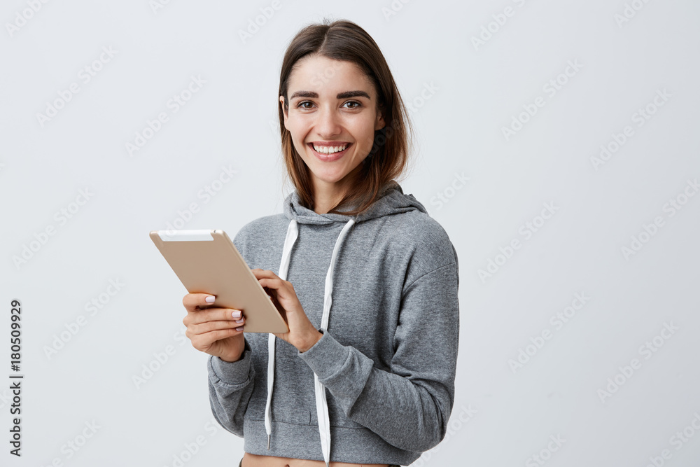 Close up of young good-looking joyful caucasian girl with dark long hair in gray sporty hoodie smiling with teeth, playing games on digital tablet, looking in camera with happy and satisfied Close up of young good-looking joyful caucasian girl with dark long hair in gray sporty hoodie smiling with teeth, playing games on digital tablet, looking in camera with happy and satisfied