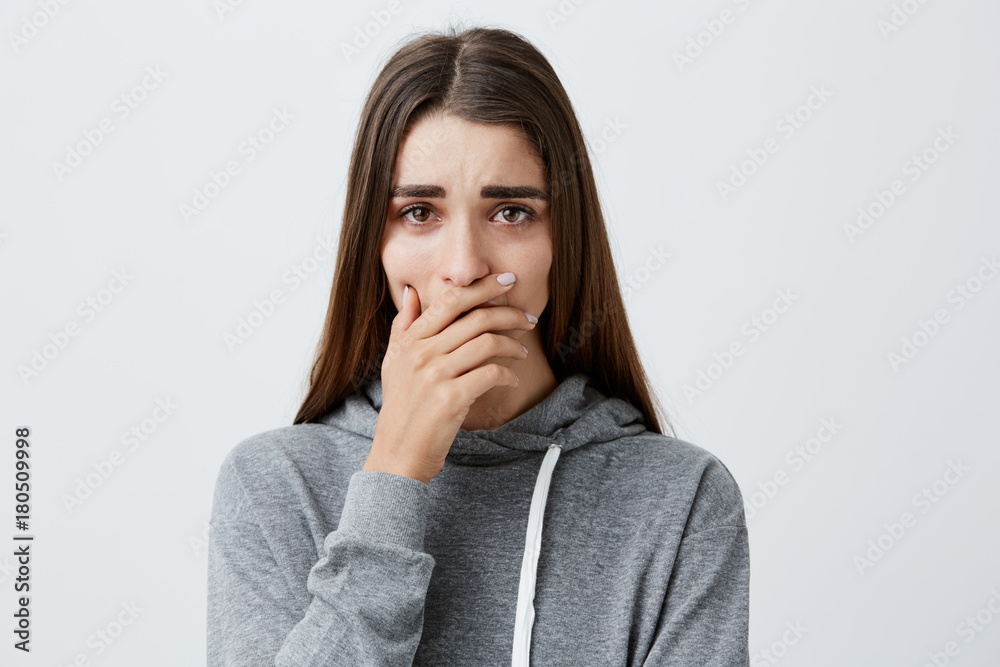 Difficult moments in life. Close up of young beautiful caucasian girl with long hair in stylish grey hoodie crying, holding hands near mouth, can't believe her beloved grandpa died.