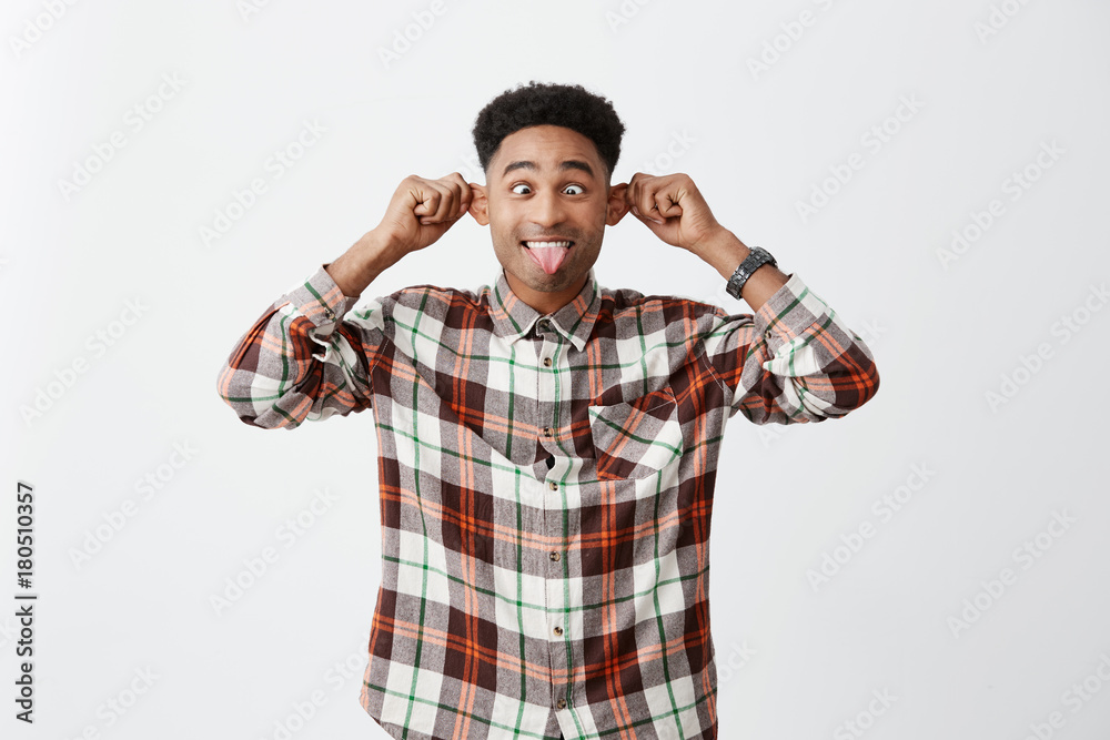 Portrait of funny beautiful young tan-skinned man with afro hairstyle in casual clothes holding ears with hands, showing tongue, mowing eyes, making silly faces, having fun. Positive emotions Portrait of funny beautiful young tan-skinned man with afro hairstyle in casual clothes holding ears with hands, showing tongue, mowing eyes, making silly faces, having fun. Positive emotions