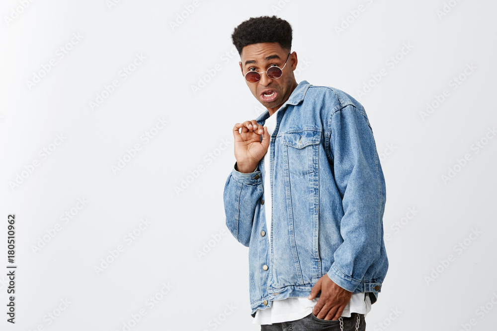 Seeing ex girlfriend with ugly guy. Portrait of young attractive  black-skinned man with afro haircut in white shirt, denim jacket and tan  glasses looking in camera with disgust expression. Stock Photo