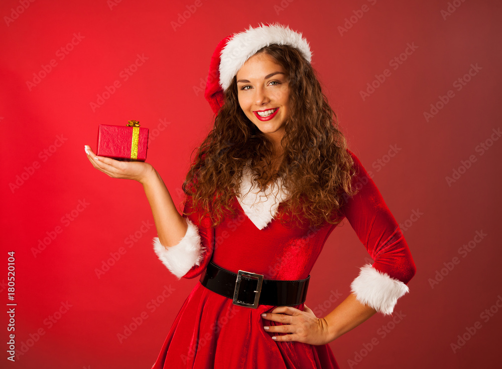 Beautiful young woman in Santa dress holds a present over red background Beautiful young woman in Santa dress holds a present over red background