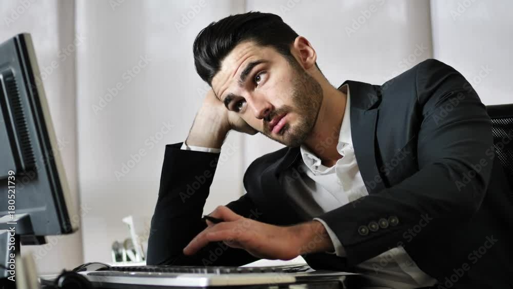 Tired bored young businessman sitting at his desk in front of his computer with his chin resting on his hand, sleepy Tired bored young businessman sitting at his desk in front of his computer with his chin resting on his hand, sleepy