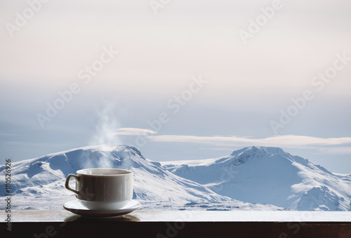 Fototapeta Cup of hot drink with steam on wooden desk and snow capped mountain view in the