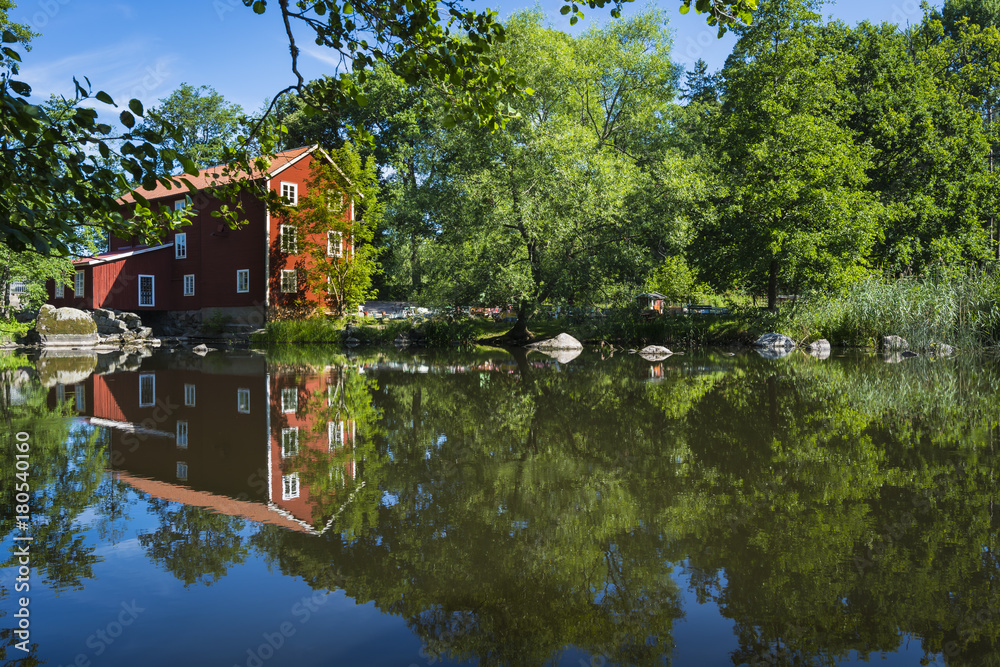 Obraz premium Beautiful picture of a Swedish watermill where the quaint café Ekenas Kvarn is situated. A colorful photo of the typical red mill with green trees and a mirror-lik pond reflecting the gorgeous surroun