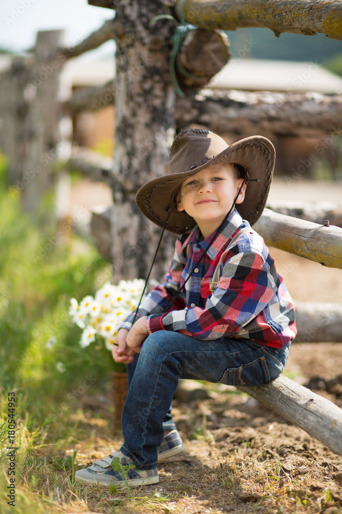 Cute fermer handsome boy cowboy in jeans enjoying summer day in village life with flowers wearing leather cow hat happyly smiling.