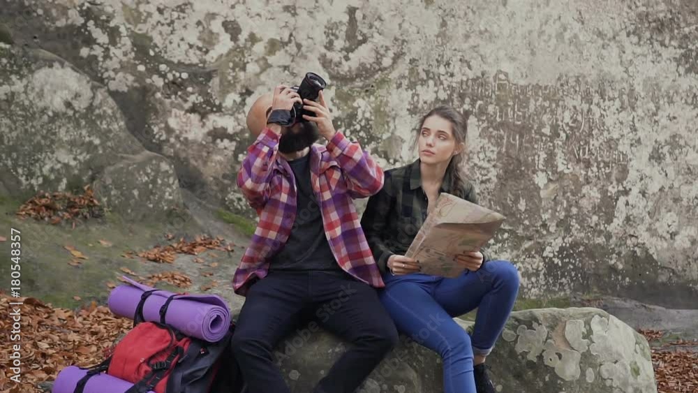 A young travel photographer with a camera, turist map and a backpack sitting with a young girl on a rock and looking at the sunset A young travel photographer with a camera, turist map and a backpack sitting with a young girl on a rock and looking at the sunset