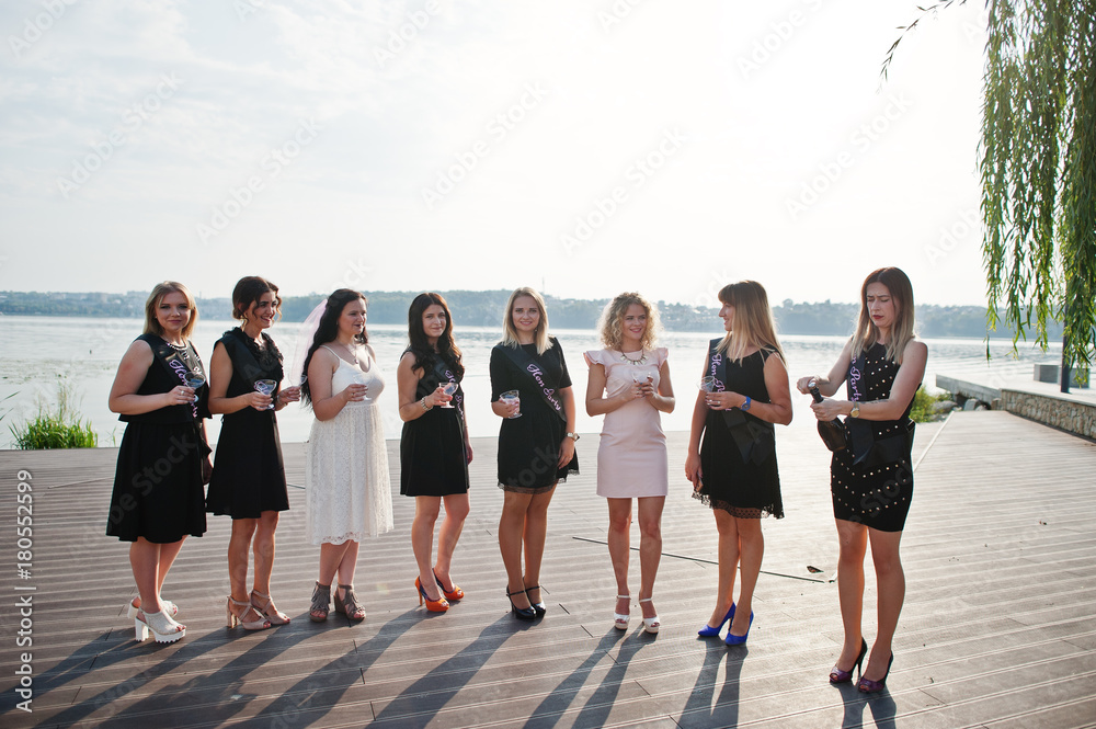 Obraz premium Group of 8 girls wear on black and 2 brides at hen party against sunny beach drinking champagne.