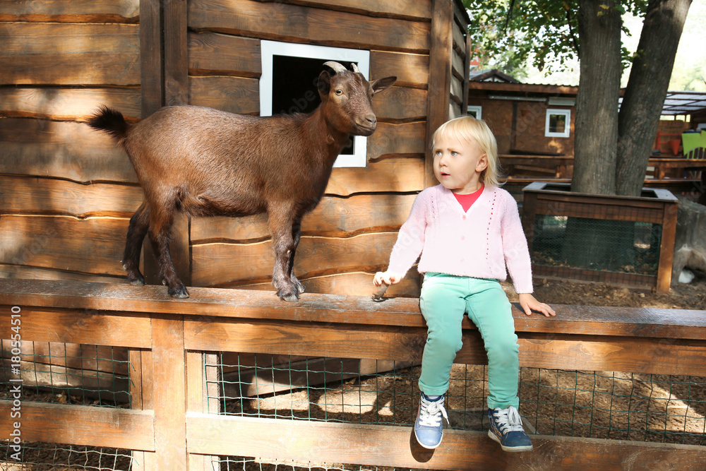 Cute little girl with funny goat sitting on wooden fence in petting zoo Cute little girl with funny goat sitting on wooden fence in petting zoo