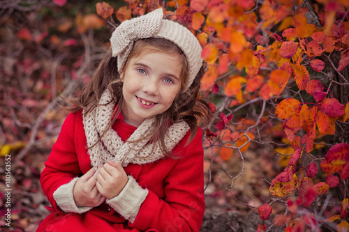 Cute young russian girl stylish dressed in warm red handmade jacket blue jeans boots and hooked headband scarf posing in autumn colorful forest pathway Face with freckling