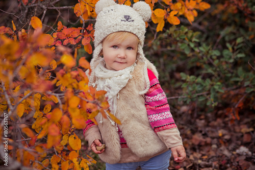 Cute young russian baby girl stylish dressed in warm white fur handmade jacket blue jeans boots and hooked hat teddy bear posing in autumn colorful forest pathway Face with pink cheek photo
