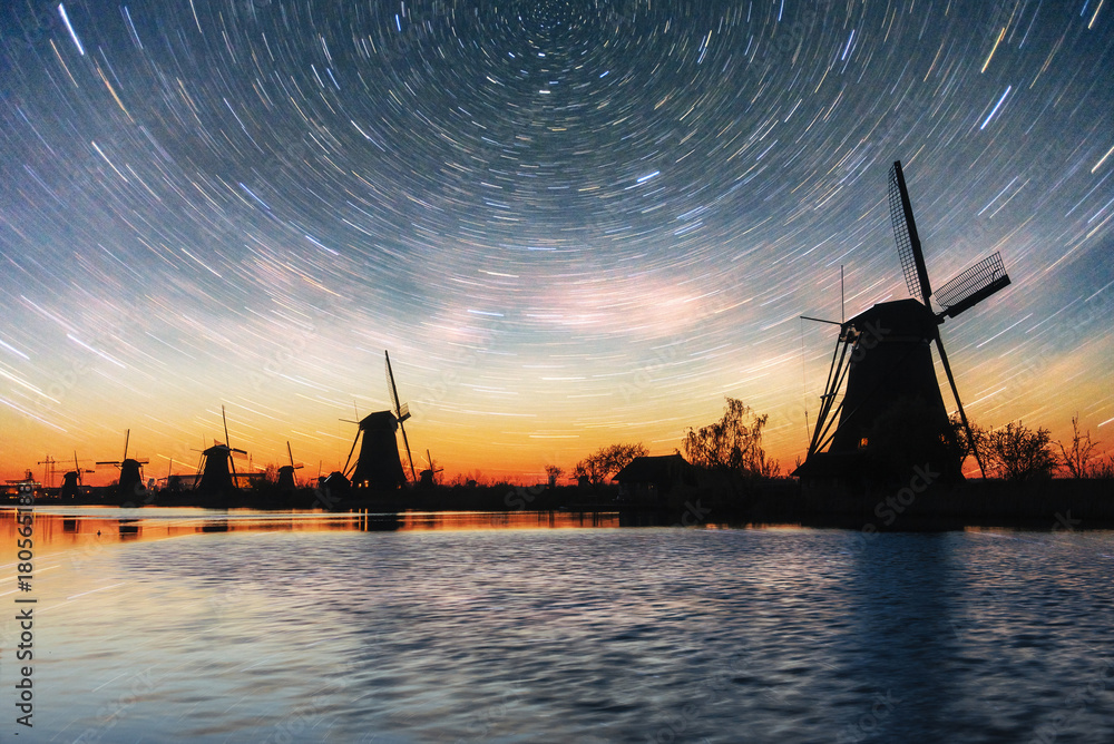 Colorful spring night with traditional Dutch windmills canal in Rotterdam. Wooden pier near the lake shore. Holland. Netherlands. Fantastic starry sky and the milky way.