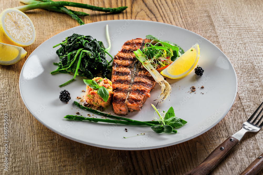 Grilled salmon fish steak with vegetable salad on a plate. Gourmet seafood meal made of fish and vegetables. Healthy food on a table. Grilled salmon fish steak with vegetable salad on a plate. Gourmet seafood meal made of fish and vegetables. Healthy food on a table.