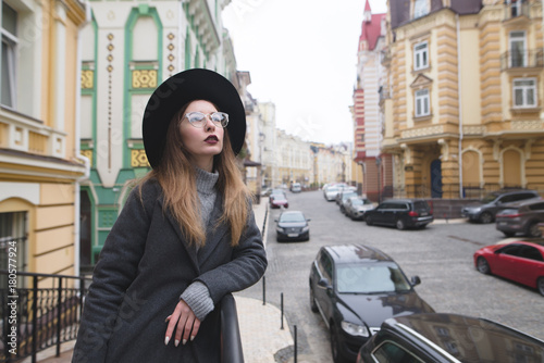 Street portrait of a stylish girl in glasses on the background of the street of the old town. Stylish hip-hop girl poses against the backdrop of a beautiful street. © bodnarphoto