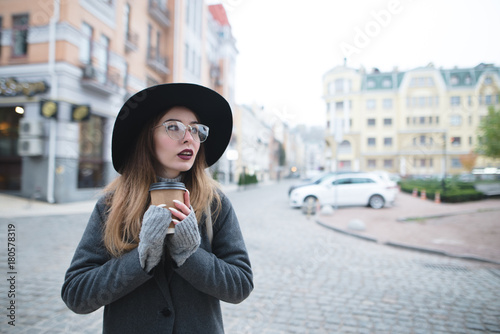 A stylish beautiful woman with coffee in her hands stands in the background of the landscape of the old city. Portrait of a girl with a cup of coffee against the background of the city. photo