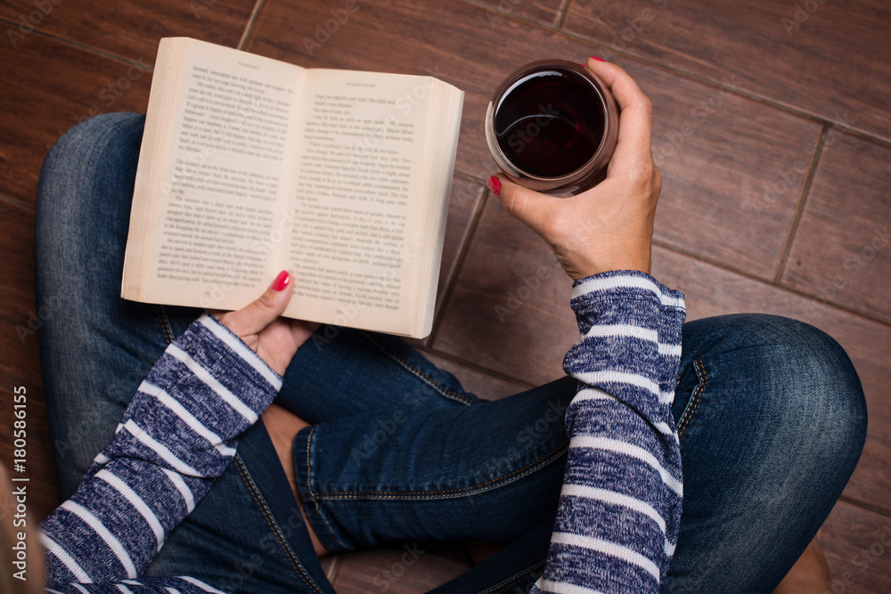 Naklejka premium Leisure, drinks, education and lifestyle concept. Woman sitting on the floor, holdind book and glass of red wine. Woman having a glass of wine and reading.
