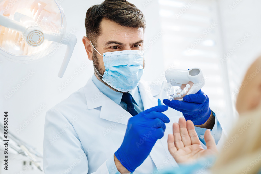 Explanation to patient. Close up of optimistic dentist with a medical mask demonstrating false teeth while looking at the patient Explanation to patient. Close up of optimistic dentist with a medical mask demonstrating false teeth while looking at the patient