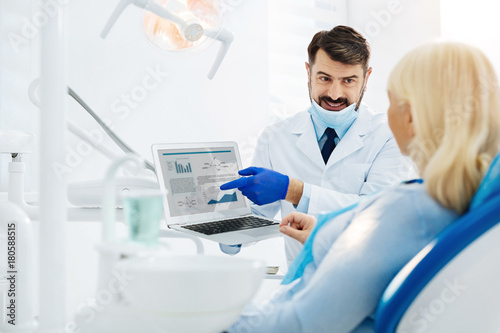 Useful information. Positive experienced dentist holding a laptop in hands while consulting the patient and having positive mood