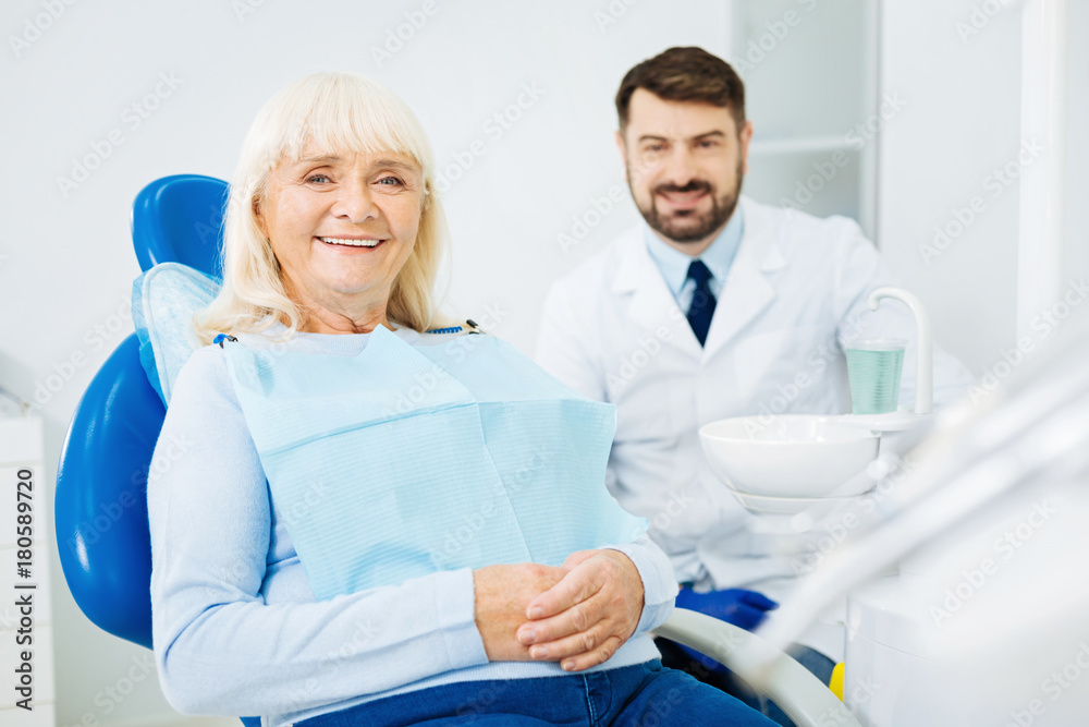 Ready for teeth cure. Waist up of cheerful patient sitting on dental chair with hands kept together while the doctor sitting in the background Ready for teeth cure. Waist up of cheerful patient sitting on dental chair with hands kept together while the doctor sitting in the background
