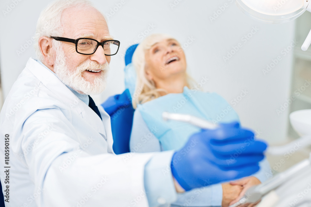 Working mood. Close up of skilled dentist holding a tool and looking away while smiling patient lying on the dental chair Working mood. Close up of skilled dentist holding a tool and looking away while smiling patient lying on the dental chair