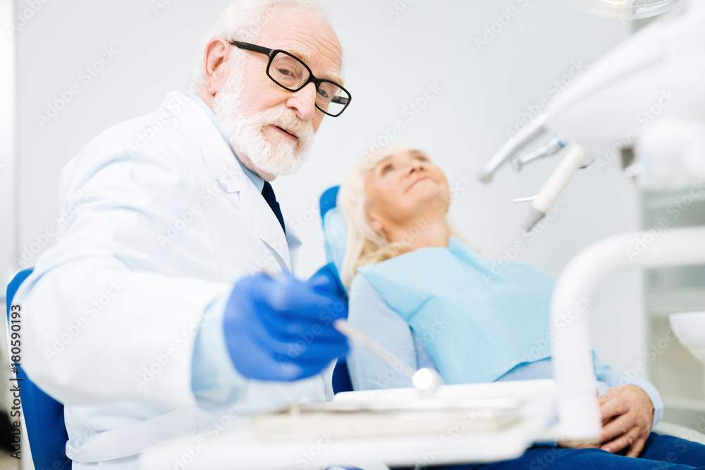 Concentration. Experienced dentist using specific tool and looking away while pleased patient lying on dental chair Concentration. Experienced dentist using specific tool and looking away while pleased patient lying on dental chair