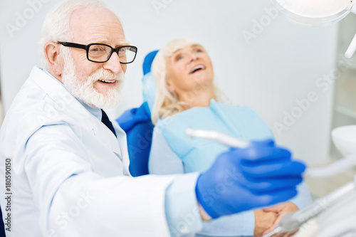 Working mood. Close up of skilled dentist holding a tool and looking away while smiling patient lying on the dental chair