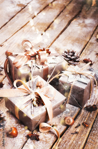 Gift boxes on old wooden background with silver stars, cones, hazelnut, anise, tape, ribbon, snowflakes. Top view, copy space, light bokeh, snow effect, toned