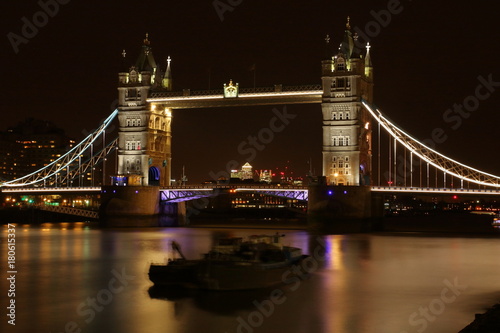 Most Tower Bridge w Londynie, Wielka Brytania, oświetlony kolorowo nocą, na Tamizie dwie niewyraźnie łodzie, barki, długie naświetlanie