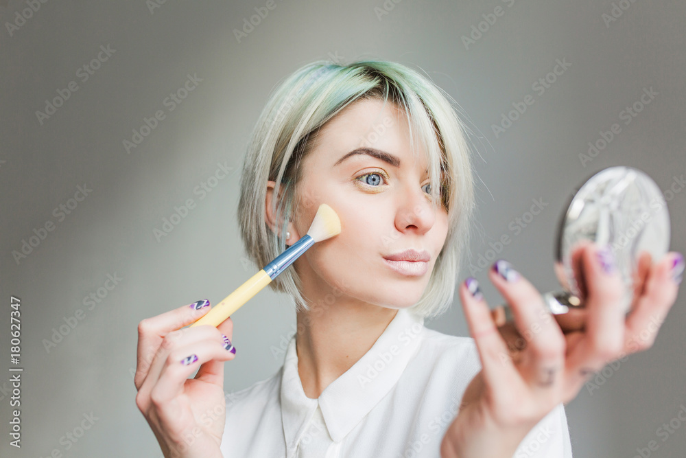 Obraz premium Close-up portrait of pretty girl with short white hair on gray background. She wears white dress, looks to the mirror in hand and powders face.