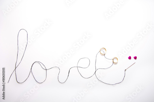 word love written with black thread and the needle inserted two gold wedding bands, there are two pink hearts and white background